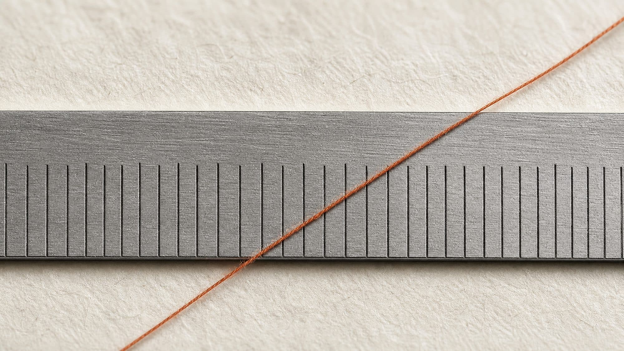 Macro photograph of an engraved steel rule on paper, a single orange thread crossing it.